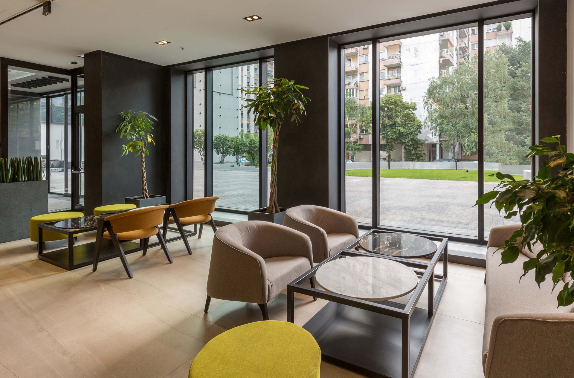 Modern lobby with beige chairs, glass tables, potted plants, and floor-to-ceiling windows overlooking a courtyard.