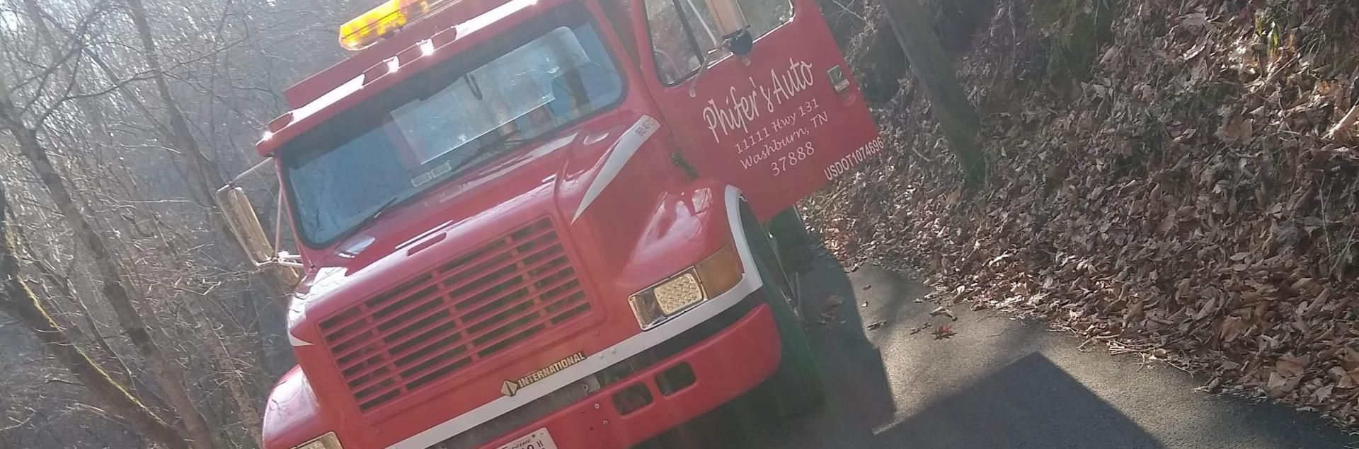 Red tow truck parked on a road with the door open. Trees and fallen leaves are visible.