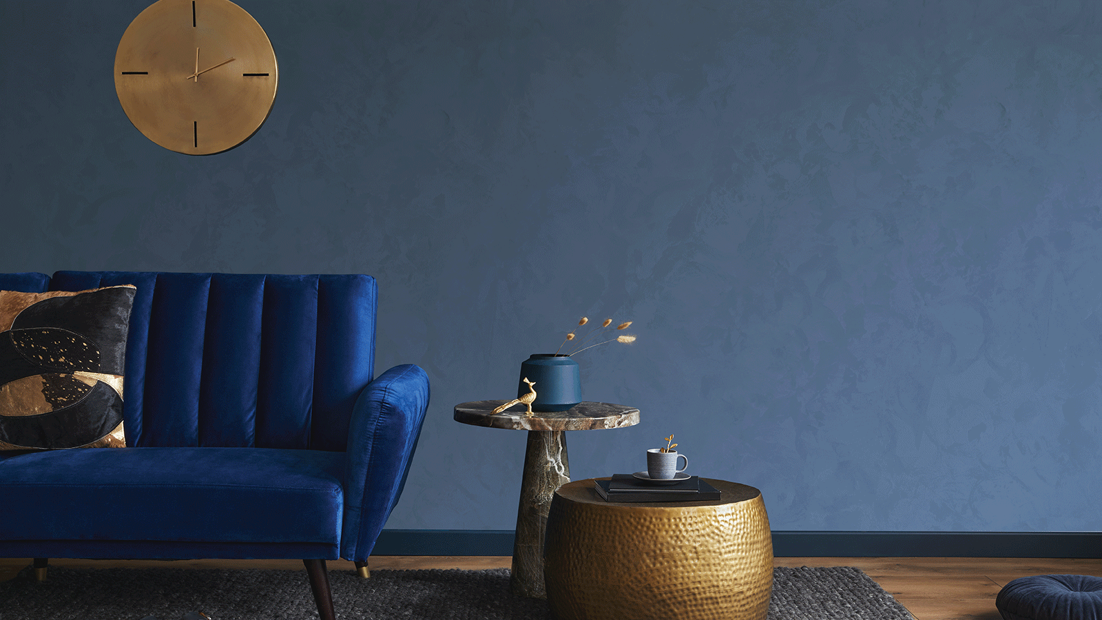 Divano in velluto blu contro una parete blu scuro, con tavolini dorati e un orologio.