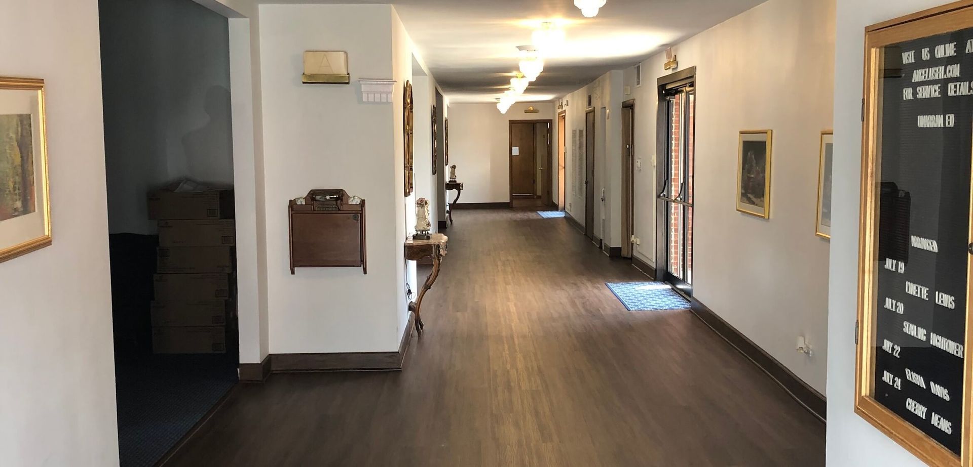 A perspective view of a long, white hallway with light wooden flooring, framed pictures on the walls, and ceiling lights.