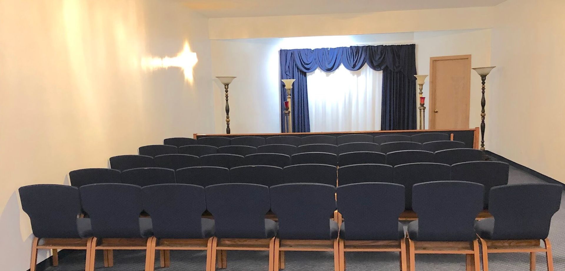 Rows of dark blue chairs face a small stage draped with navy blue curtains in a brightly lit auditorium.