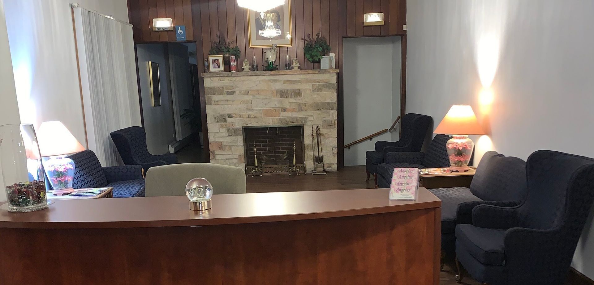 A lobby or waiting area features a front desk, a stone fireplace, wood-paneled walls, and several dark blue armchairs.