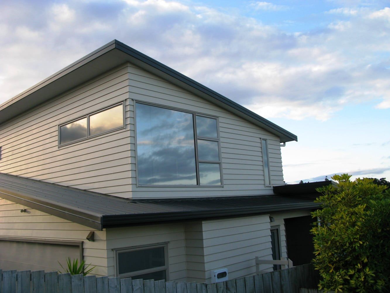 Two-story house with white siding and large windows, reflecting a cloudy sky.