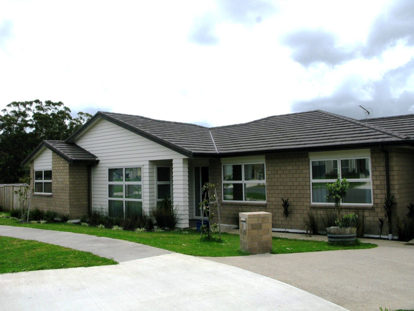 A single-story brick house with white siding and a gray roof, driveway, and small front yard under a cloudy sky.