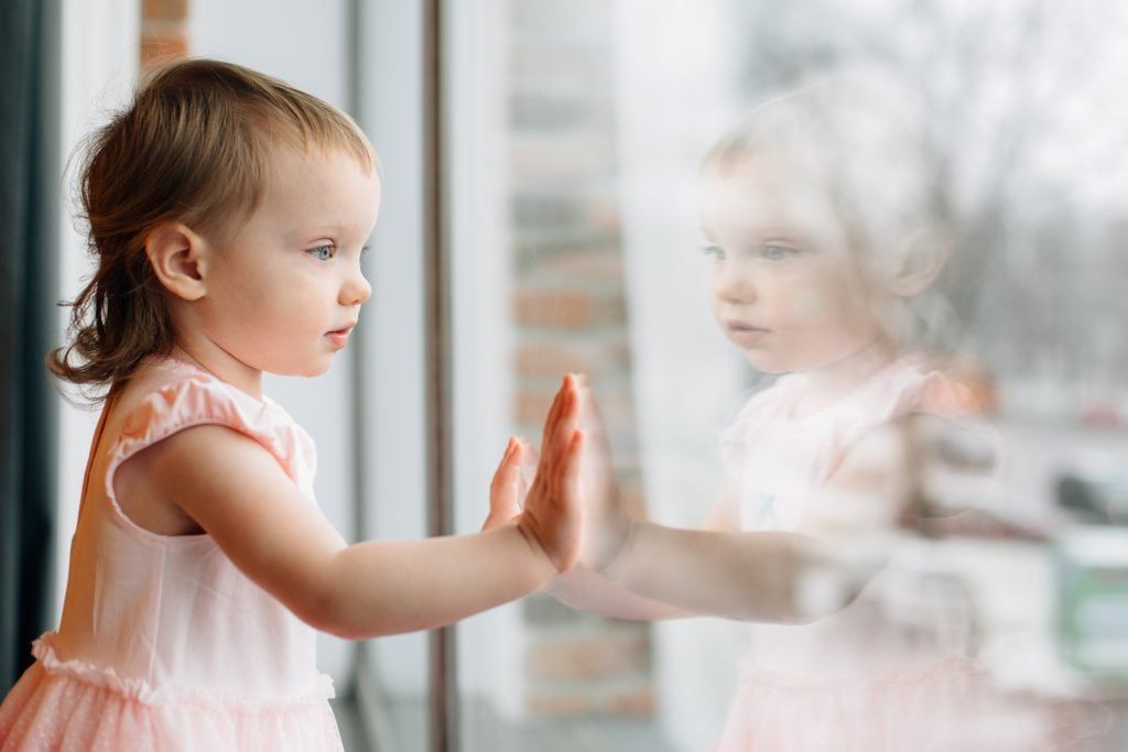 Child in a pink dress, touching a window. The child's reflection is also visible.