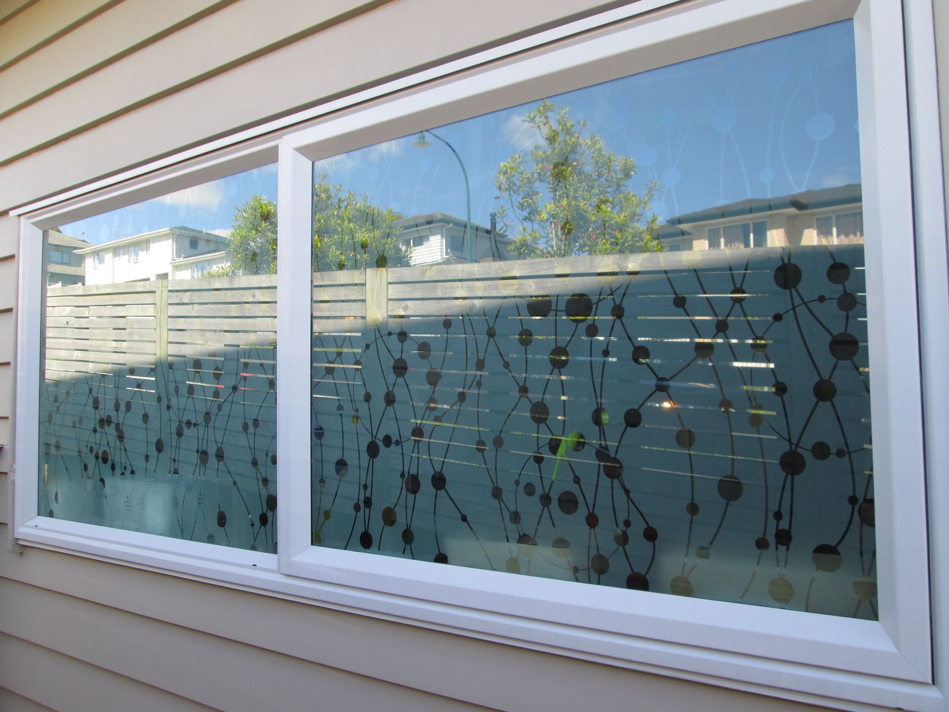 White-framed window with decorative pattern, reflecting a neighborhood. Brown siding.