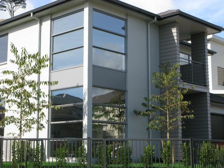 Two-story modern house with large windows reflecting the sky, gray exterior, small trees in front of a black fence.