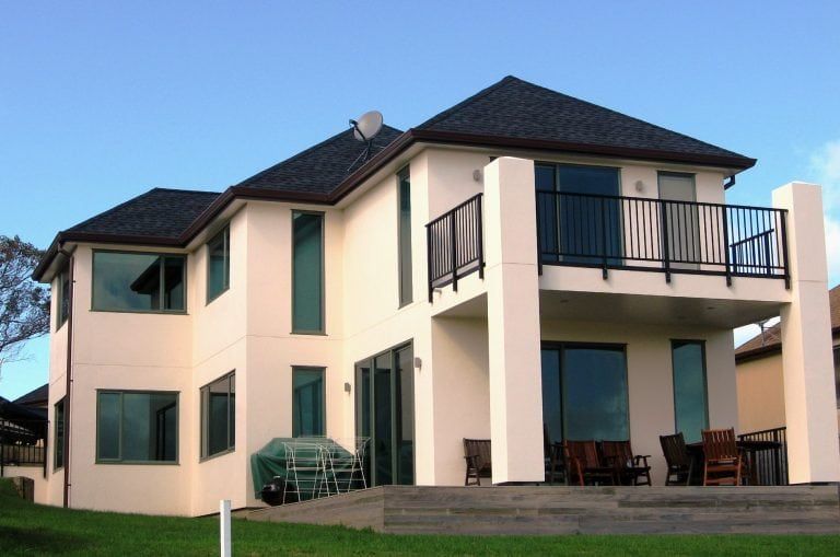 Two-story cream-colored house with black roof, balcony, and large windows on a bright day.