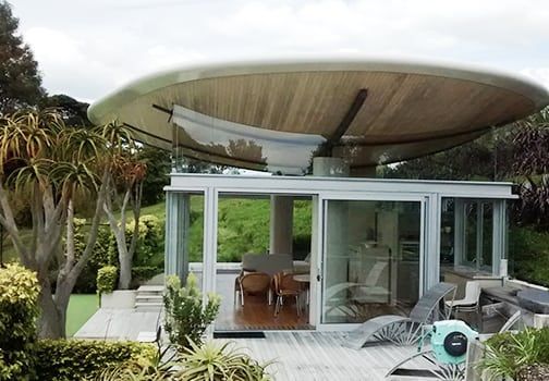 Modern house with a large, circular roof. Glass walls reveal a deck, furniture, and green landscape.