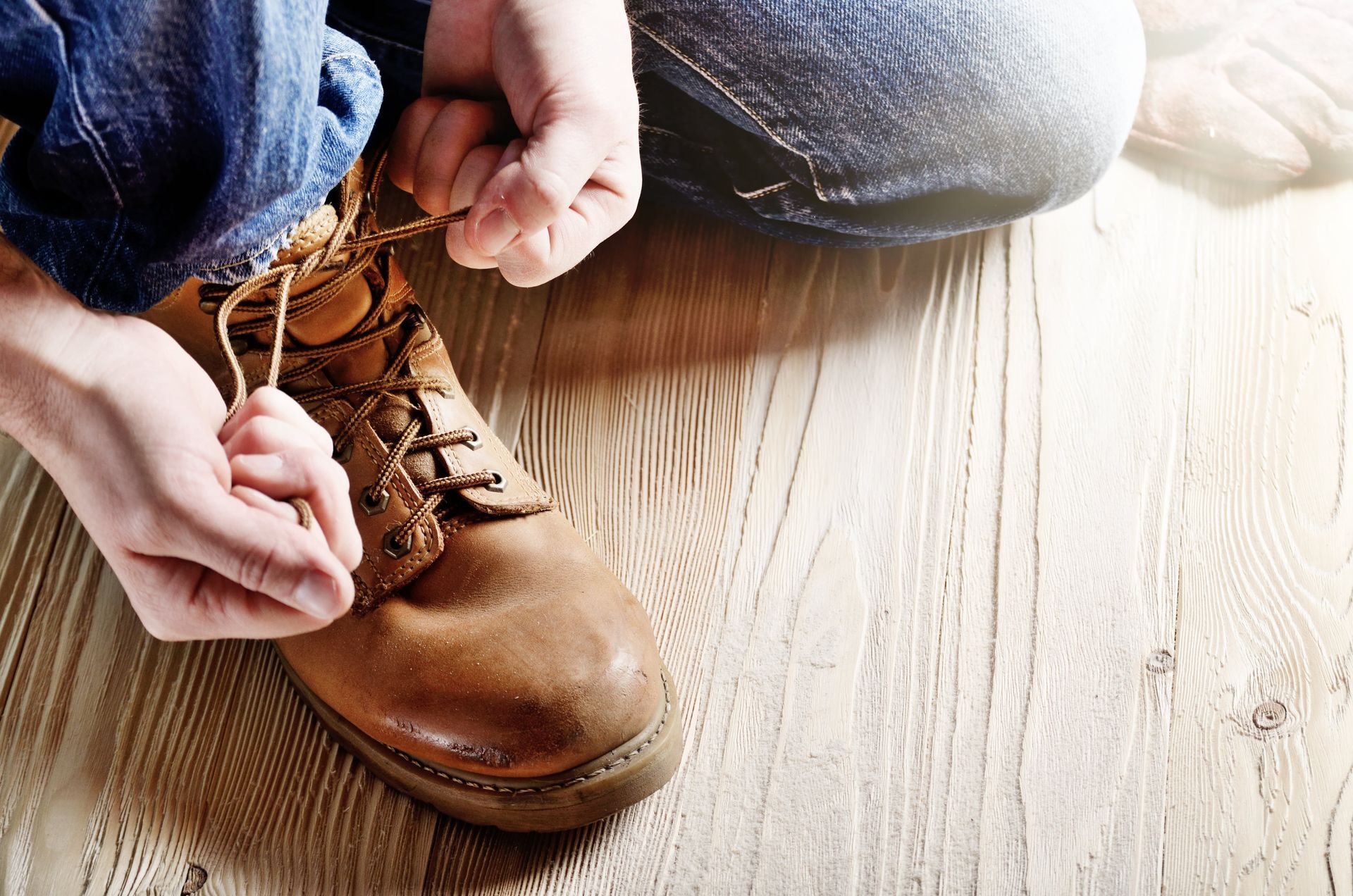 Person tying the shoelaces of a brown leather boot on a wooden floor.