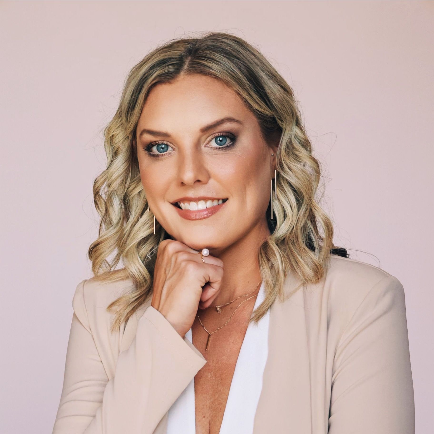 A woman with long blonde hair and blue eyes is wearing a tan jacket.