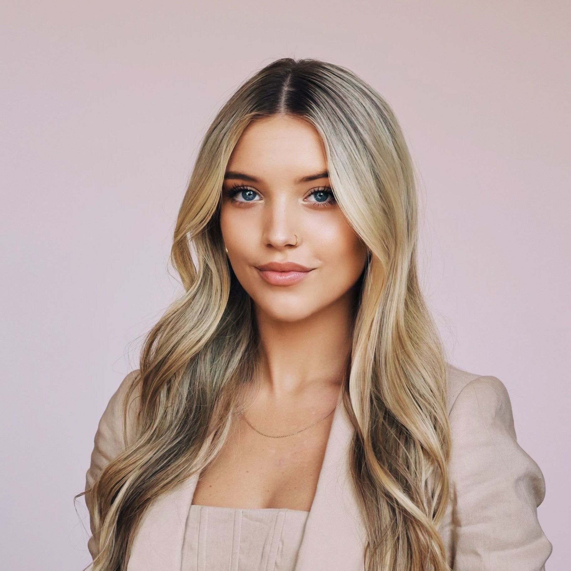 A woman with long blonde hair and blue eyes is wearing a tan jacket.