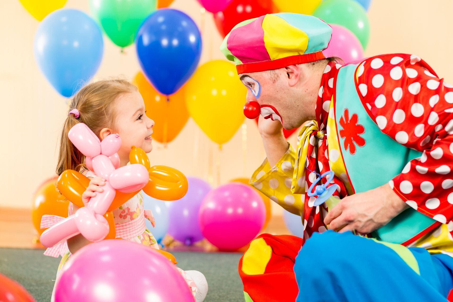 A clown is playing with a little girl in a room filled with balloons.