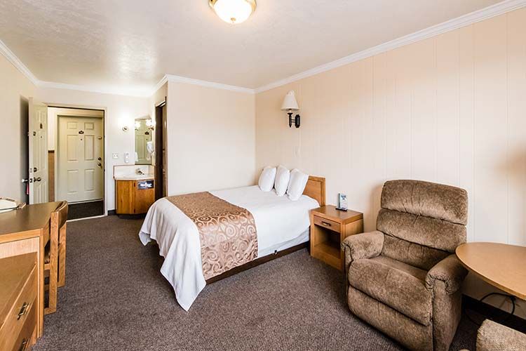 Motel room with bed, recliner, desk, and bathroom doorway. Beige walls and brown carpet.