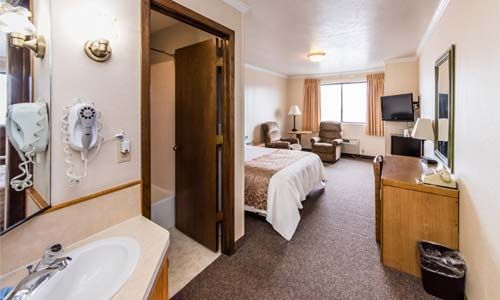Hotel room interior with a bed, desk, bathroom entrance, and recliner. Brown carpet, beige walls.