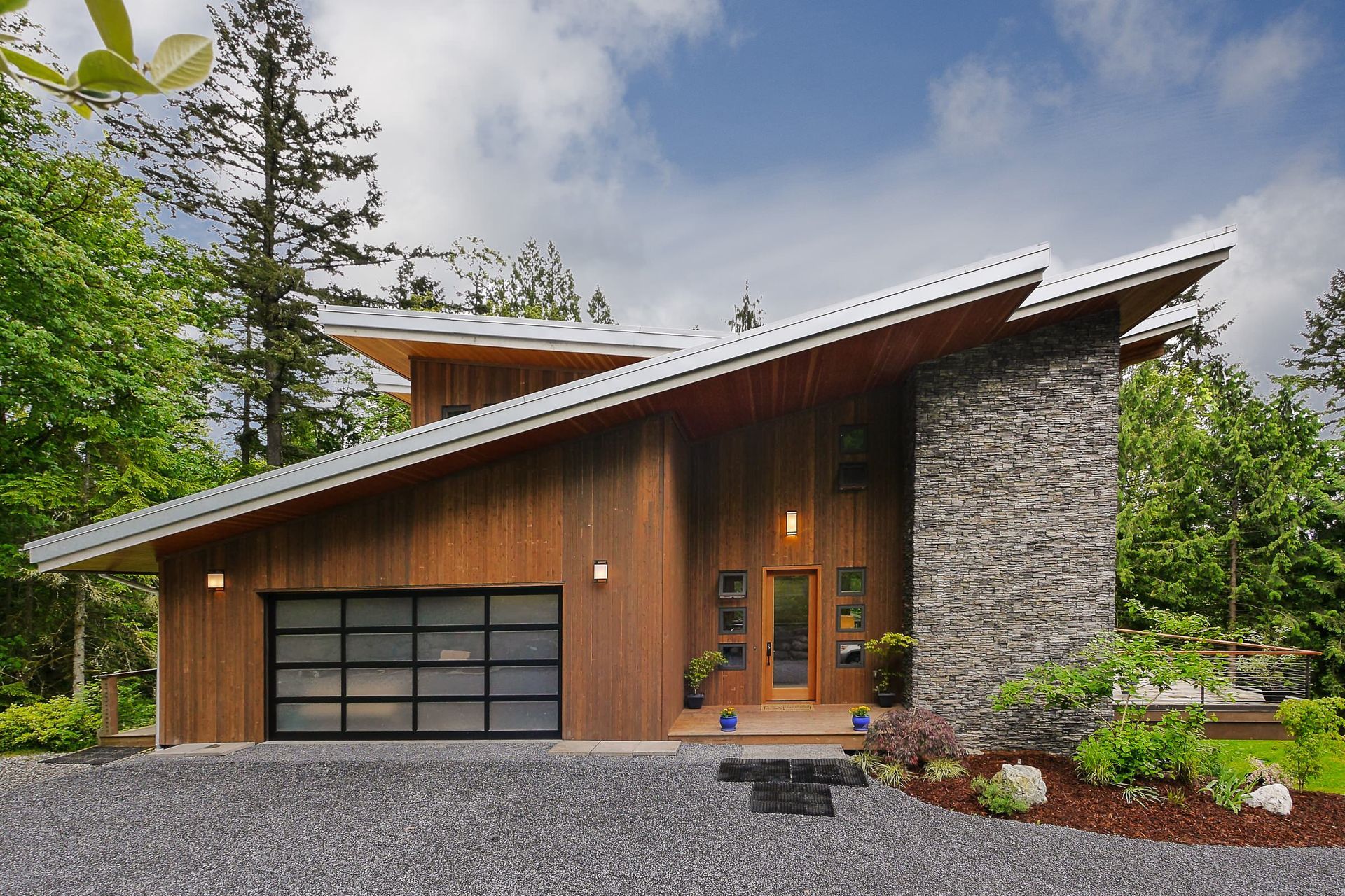 Modern house with slanted roof, wood siding, stone chimney, and glass garage door. Gravel driveway, trees.