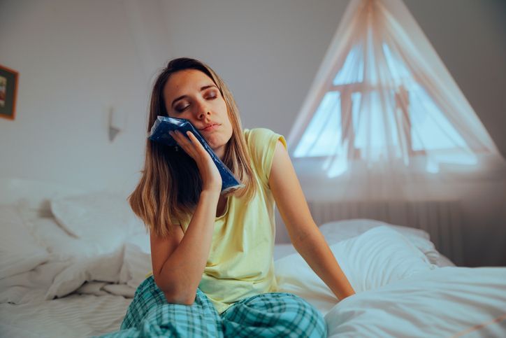 Woman sitting on a bed, holding a blue cloth to her face in a softly lit bedroom.