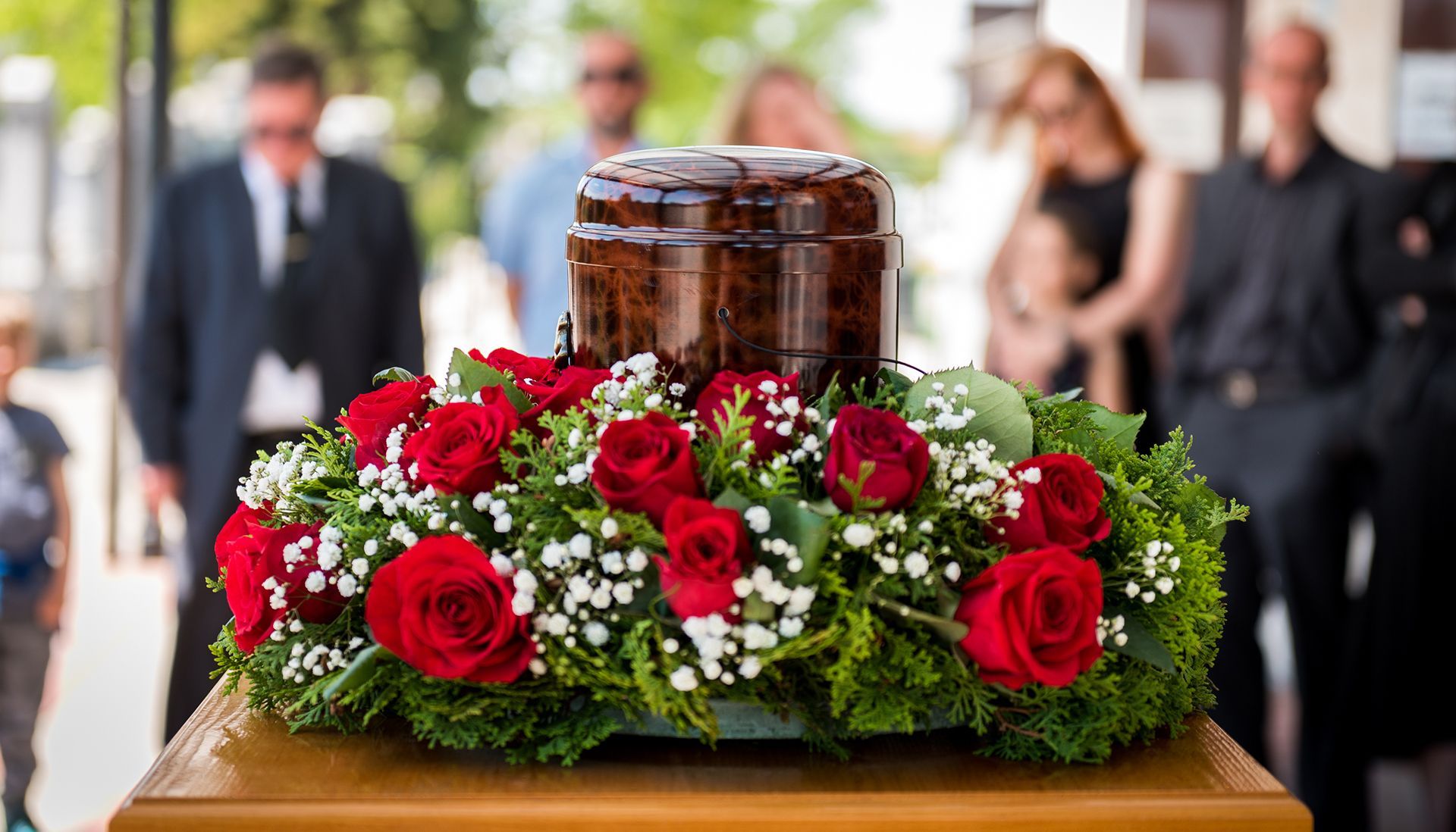 A urn surrounded by red roses and baby 's breath is on a coffin at a funeral.