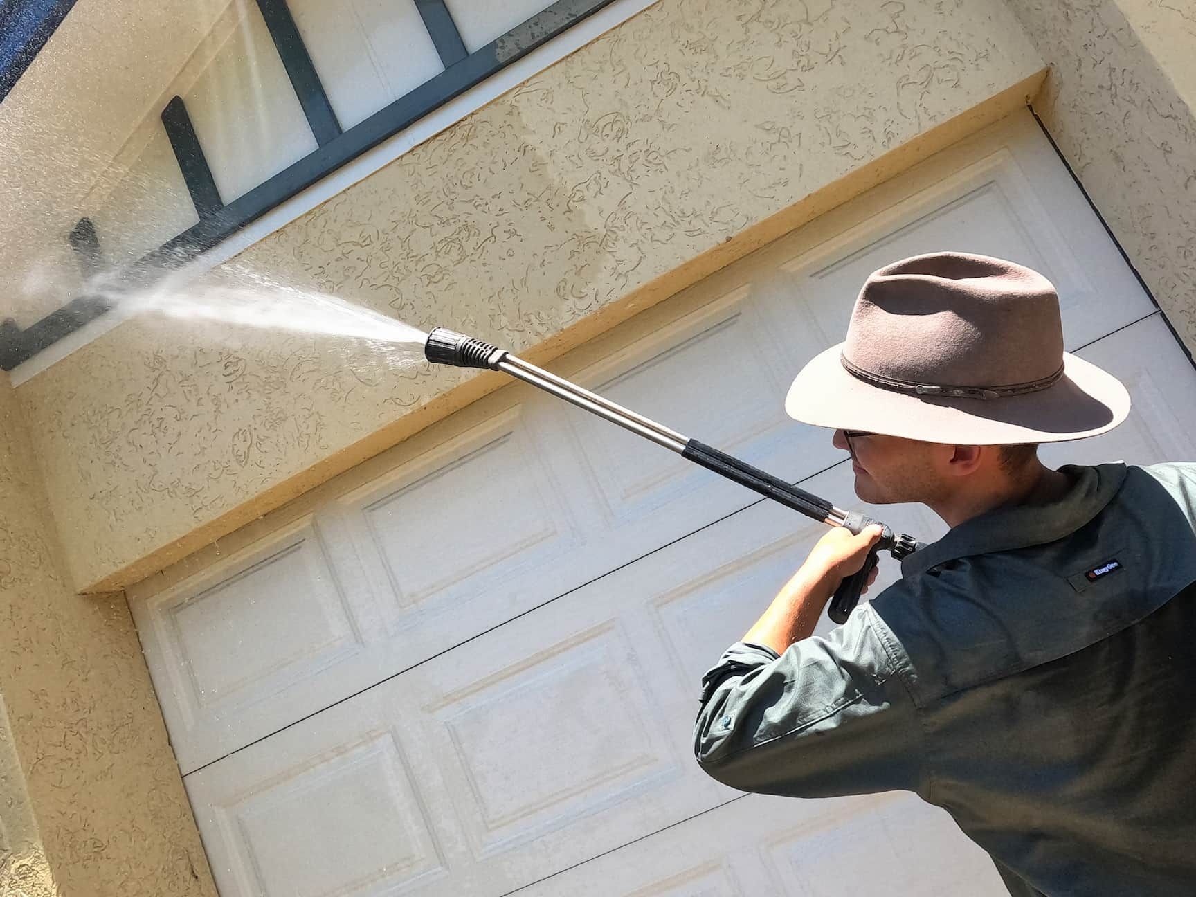A Man in A Hat Using a High Pressure Washer to Clean a Garage Door — Zac's Bin and Pressure Cleaning In Parramatta Park, QLD