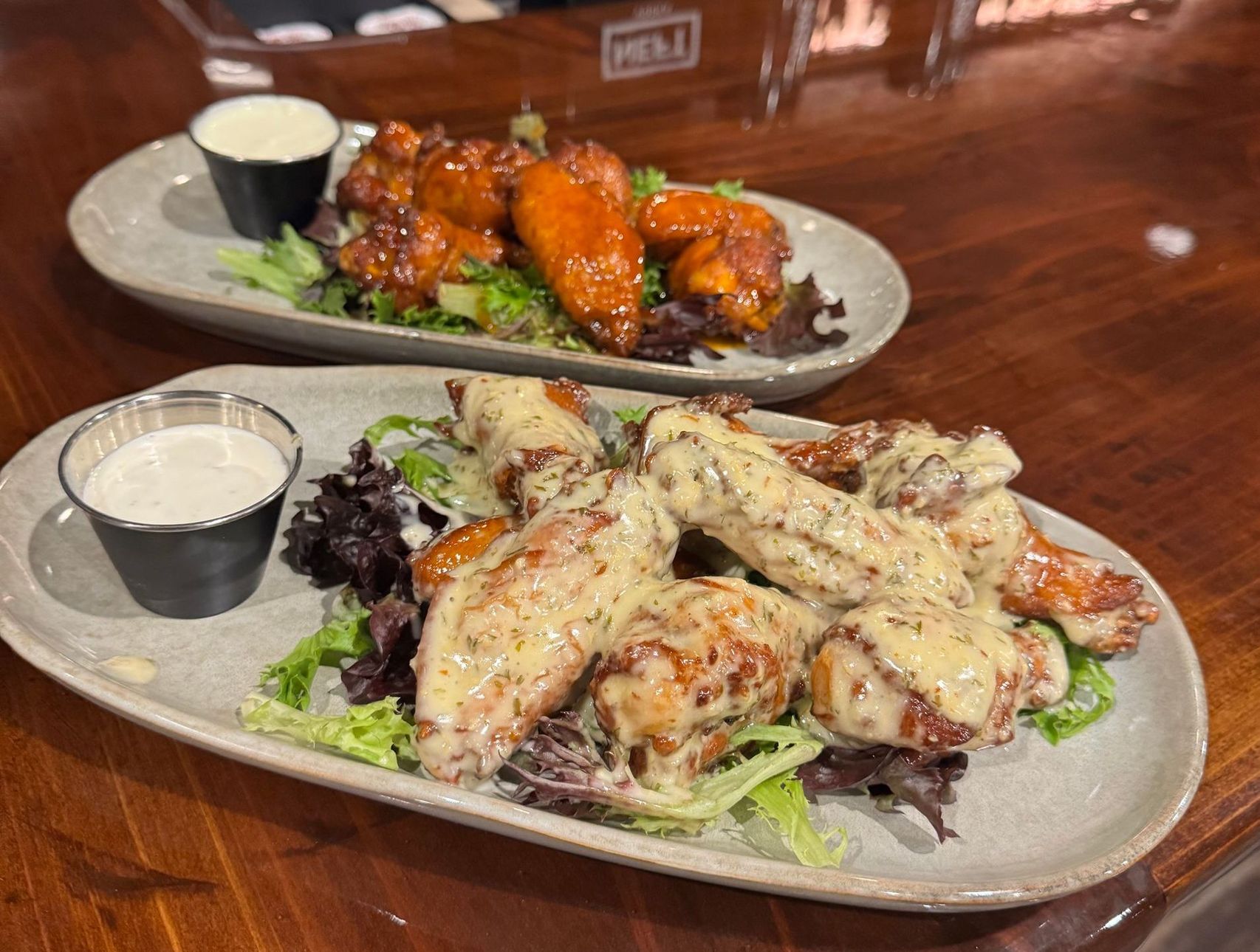 Two plates of chicken wings served on a wooden table, one with a red sauce and one with a creamy garlic sauce.