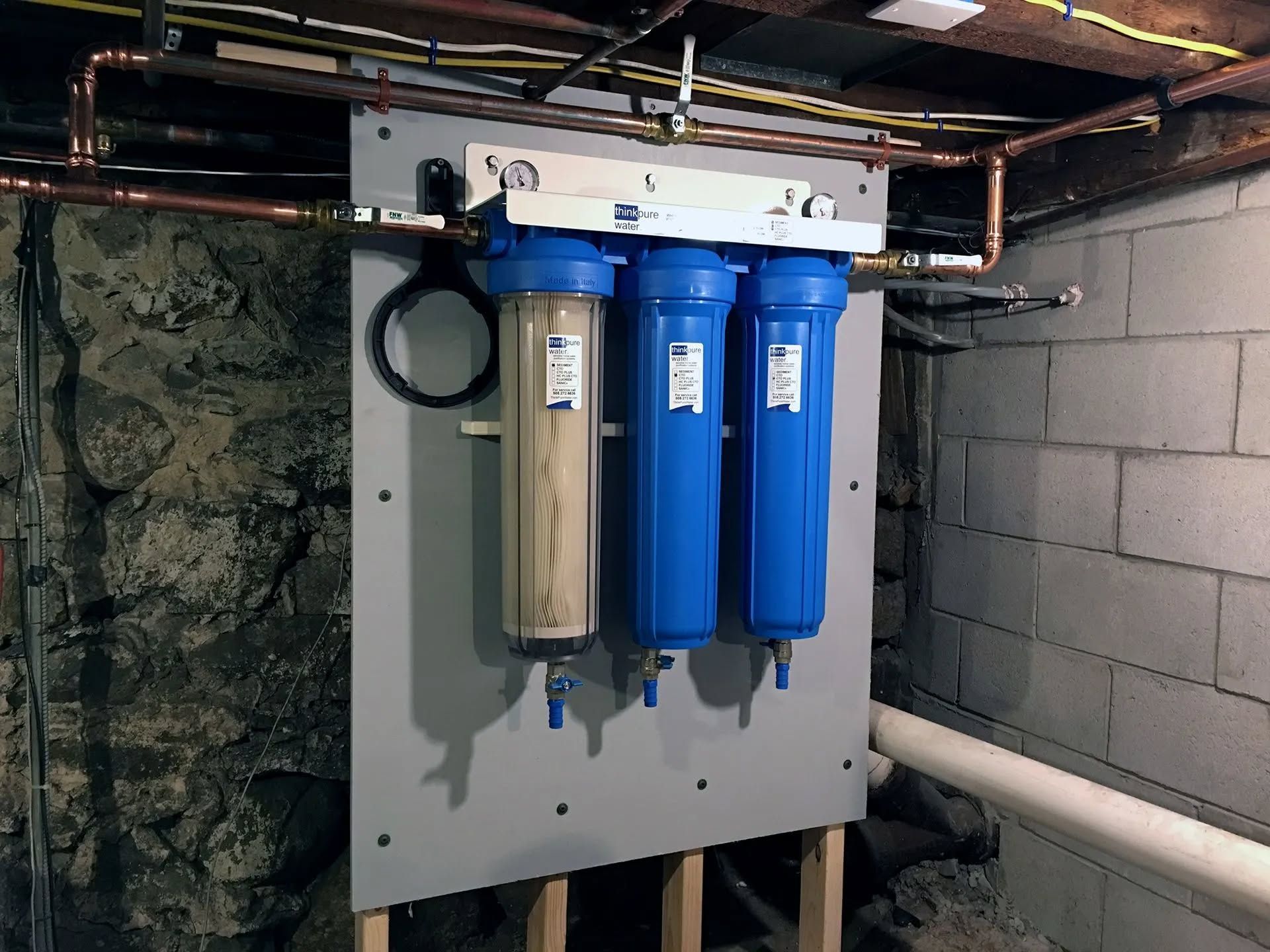 Wall-mounted water filtration system with three blue canisters in a basement utility area