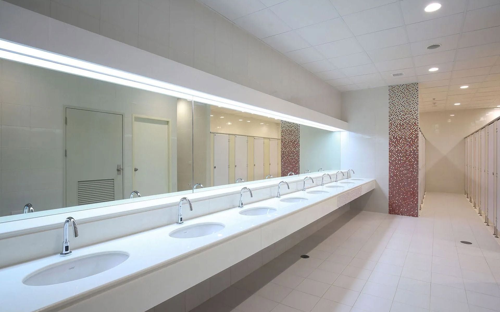 Bright public restroom with long white sink counter, mirrors, and stalls along the wall