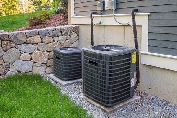 Two outdoor air conditioner units beside a house, next to a stone retaining wall and grass.