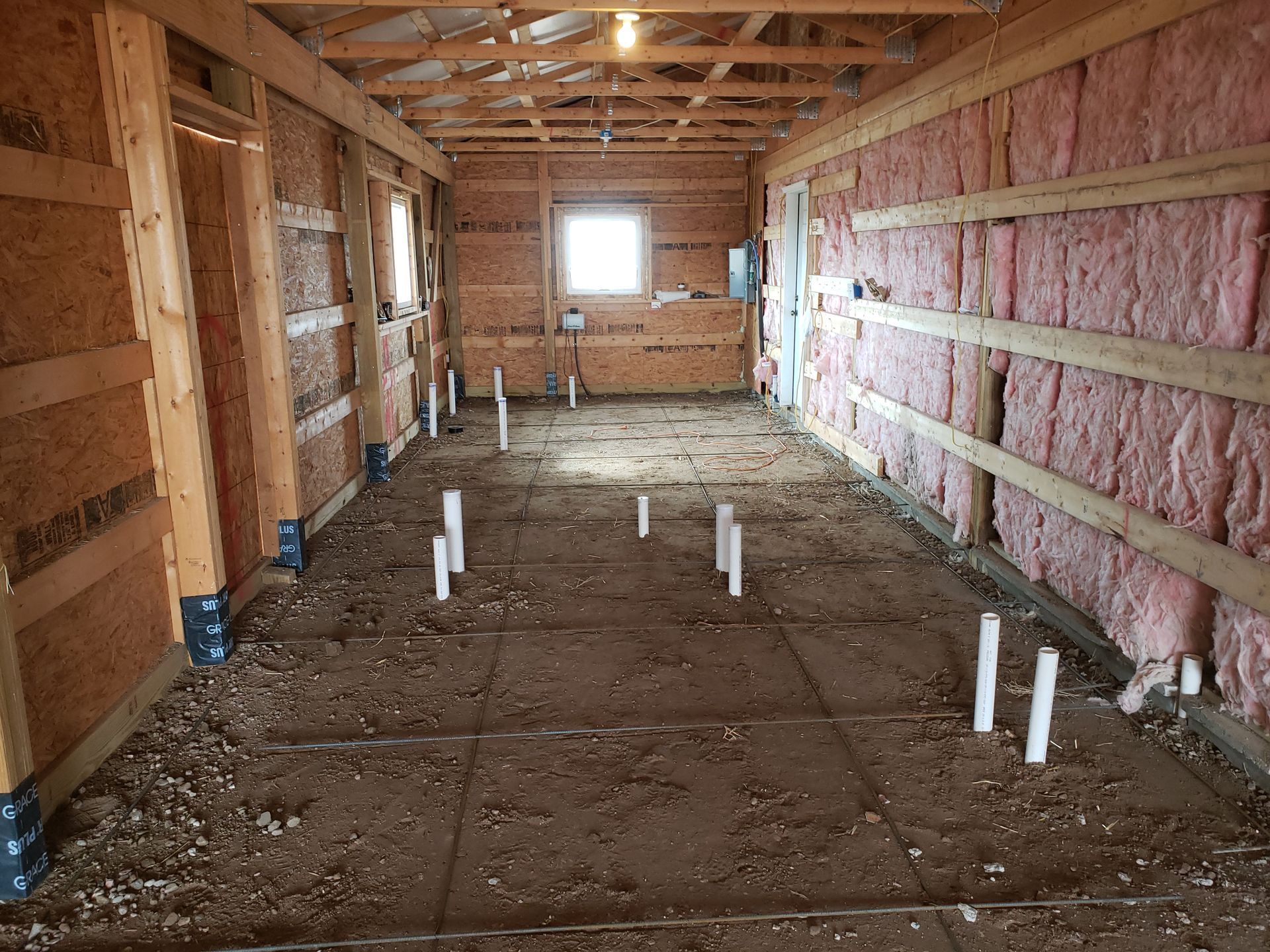 Interior of an unfinished wooden house with exposed framing, pink insulation, and PVC pipes on the floor