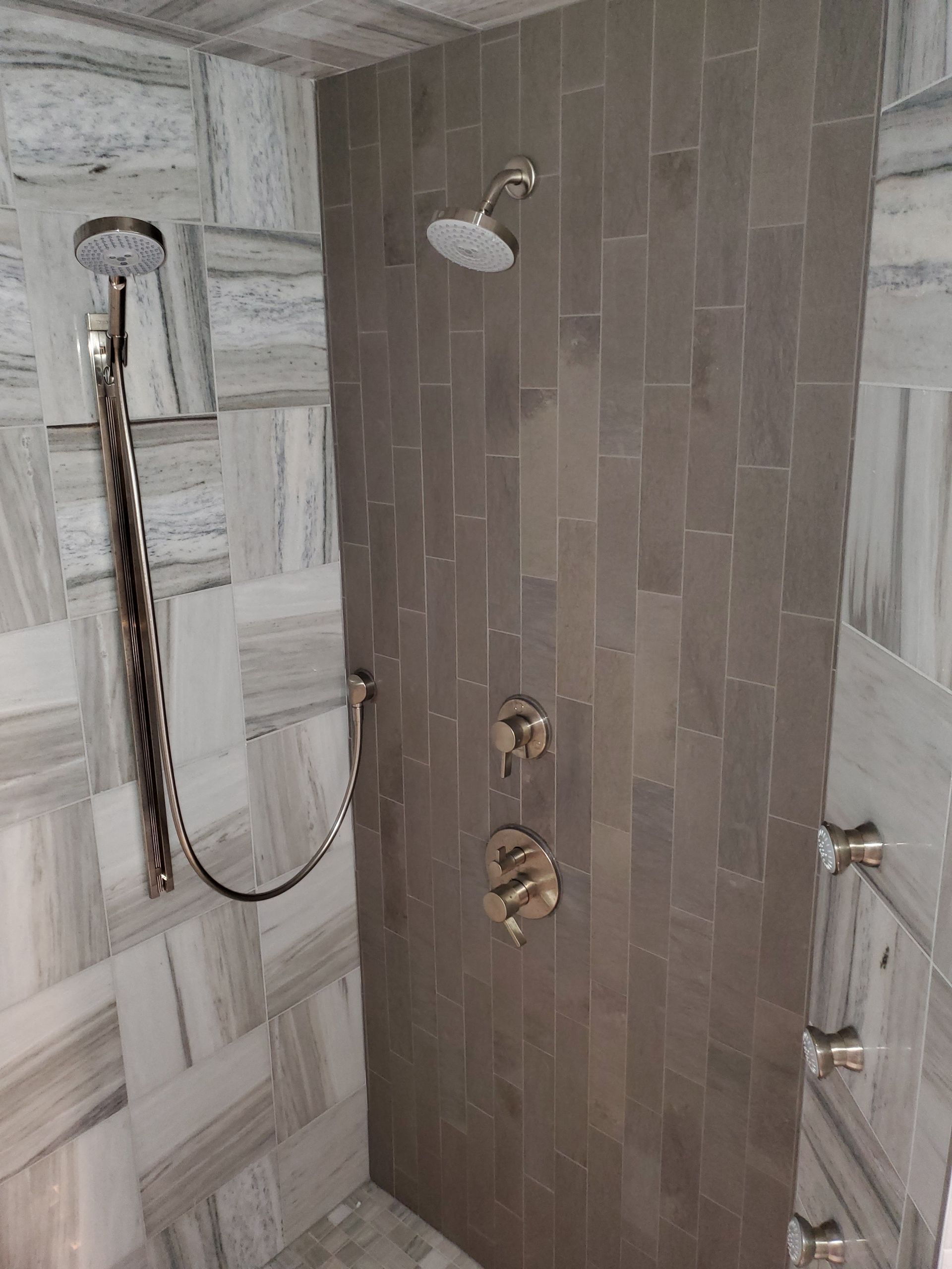 Small tiled shower with handheld and overhead showerheads, gray and white stone walls.