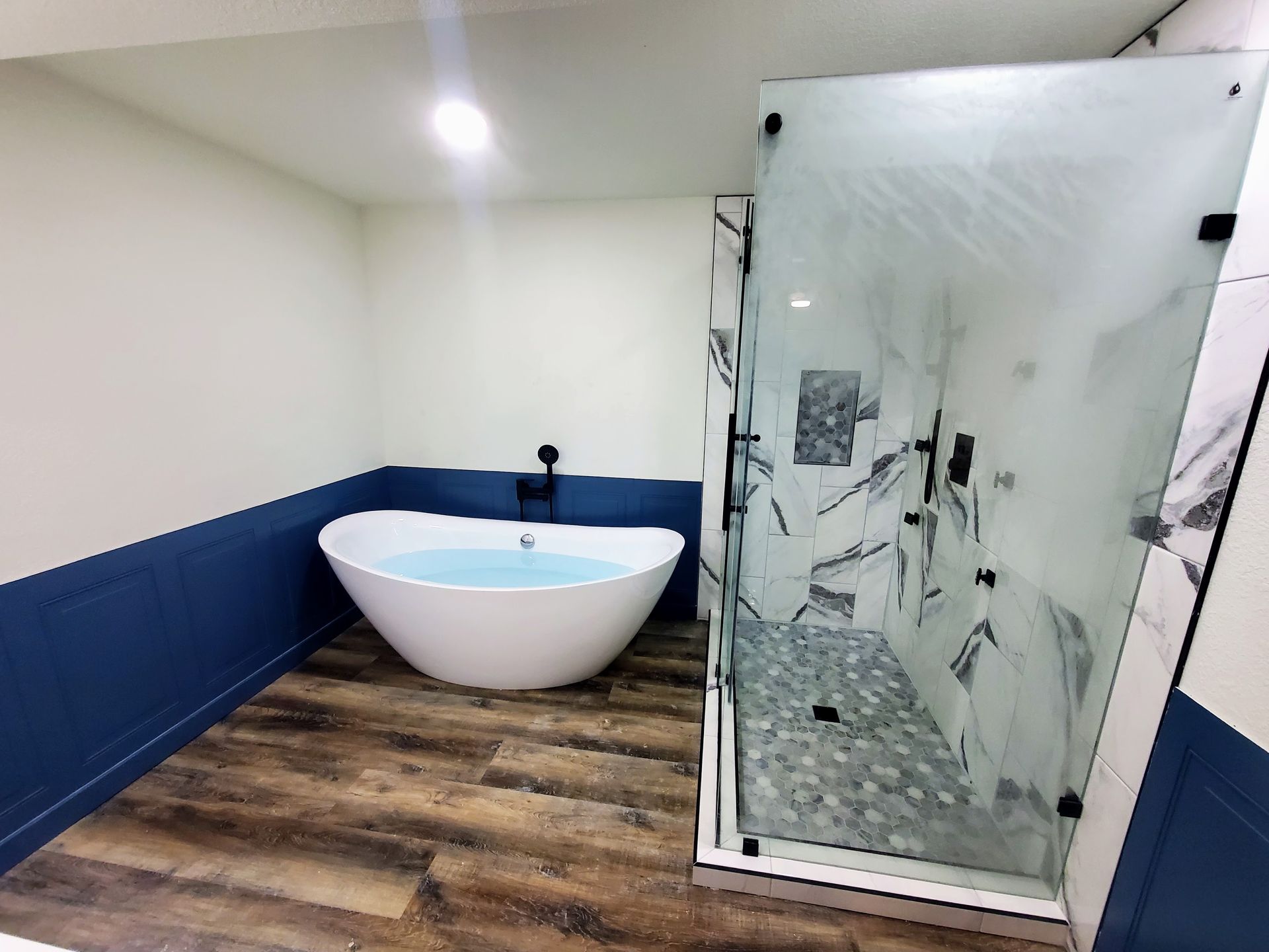 Modern bathroom with white tub, glass shower, blue wainscoting, and wood-look flooring
