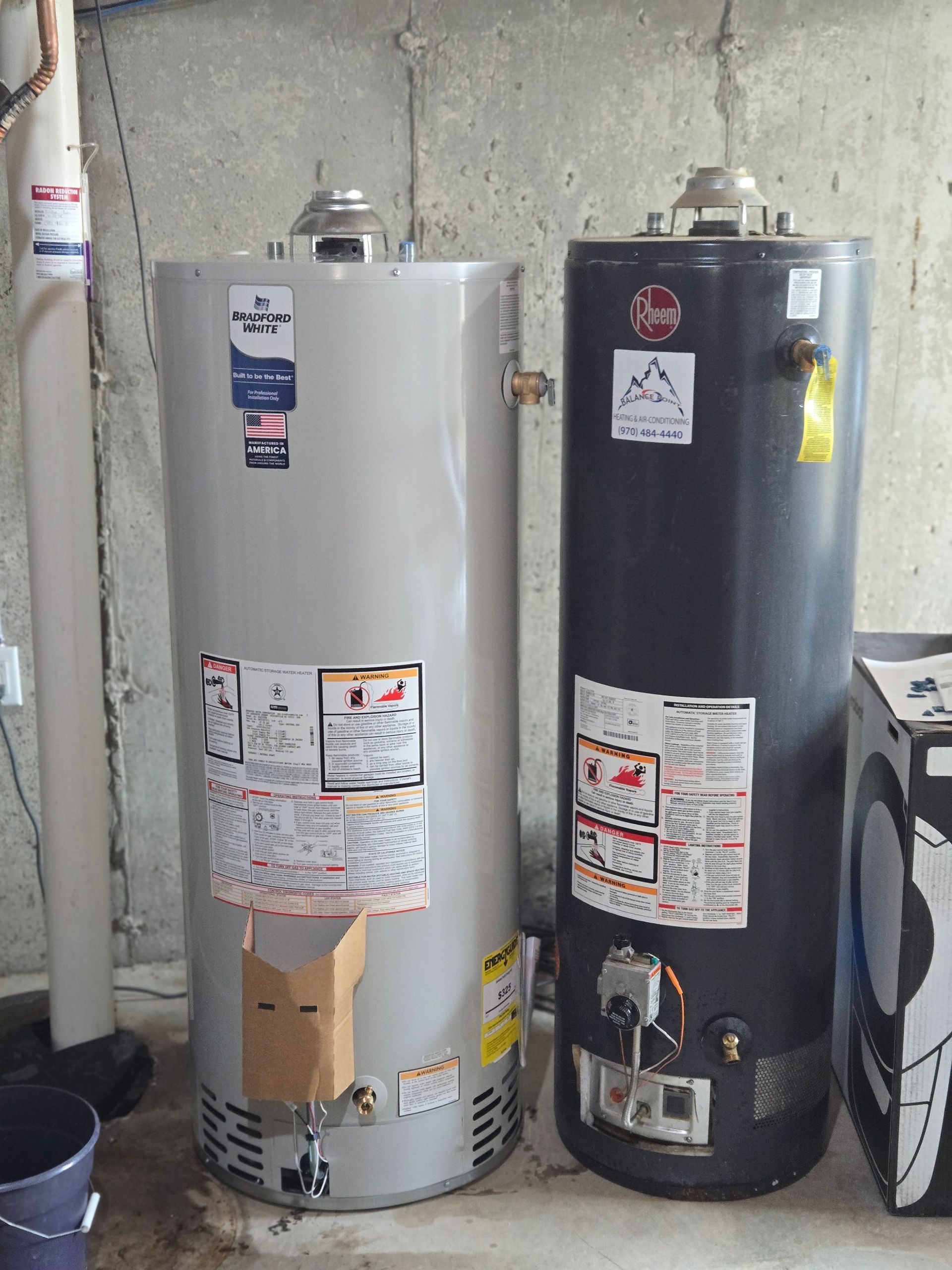 Two water heaters in a utility room, one white and one dark gray, with pipes and labels.