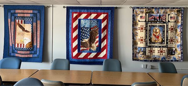 A room with tables and chairs and three quilts hanging on the wall.