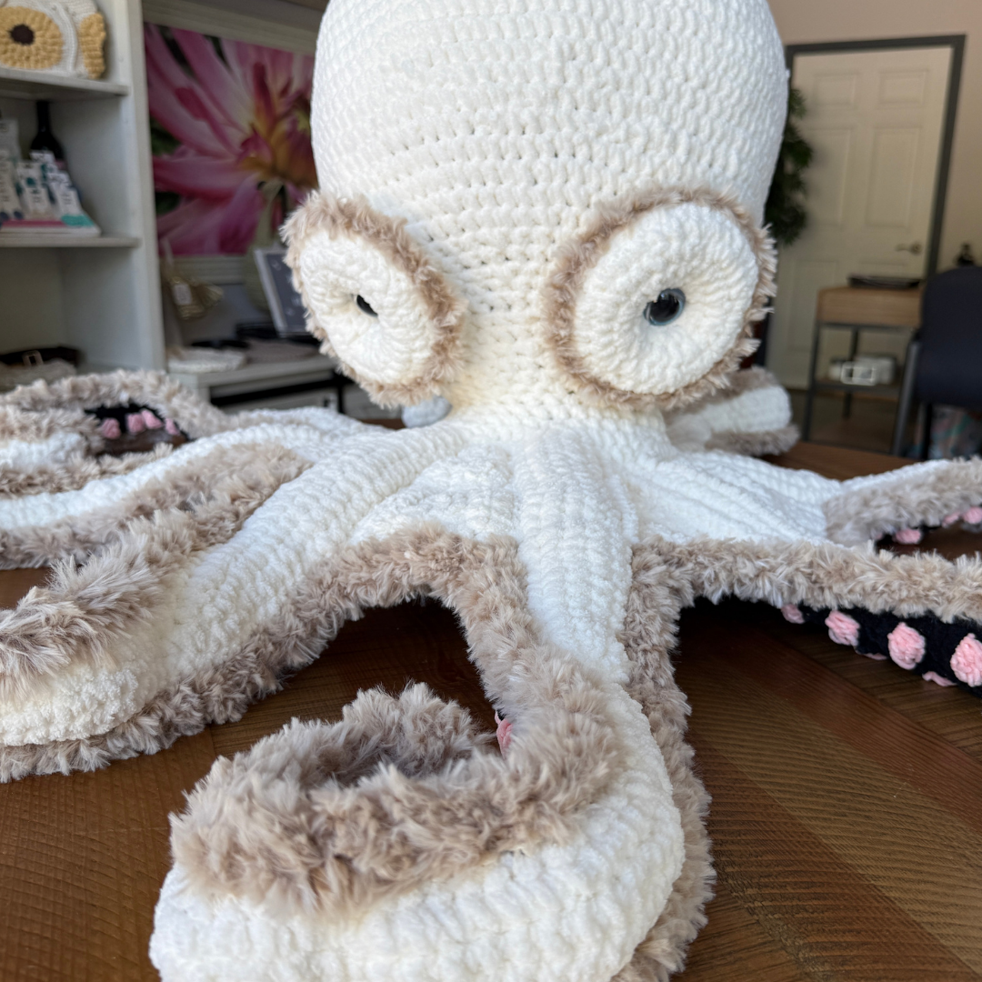 Crocheted white octopus with beige and pink tentacles, large googly eyes, sitting on a table.