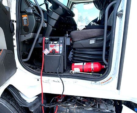 Interior of a semi-truck cab with dashboard, seat, and fire extinguisher. Black and red electronics.