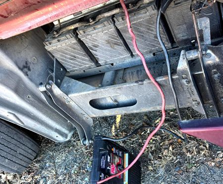 Three car batteries in a vehicle, connected to a charger with red and black cables.