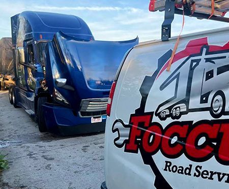 Blue semi-truck with open hood next to a white service van with a logo that says “Focus Road Service”.