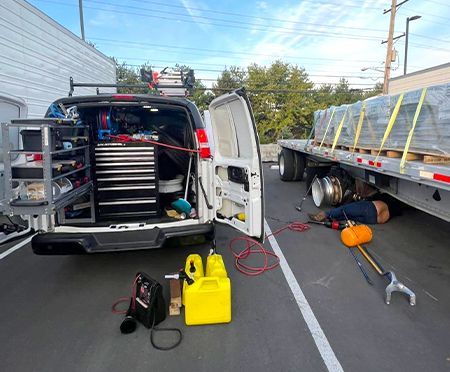 Mechanic working on truck wheel. Van with tools open, flatbed trailer with pallets.