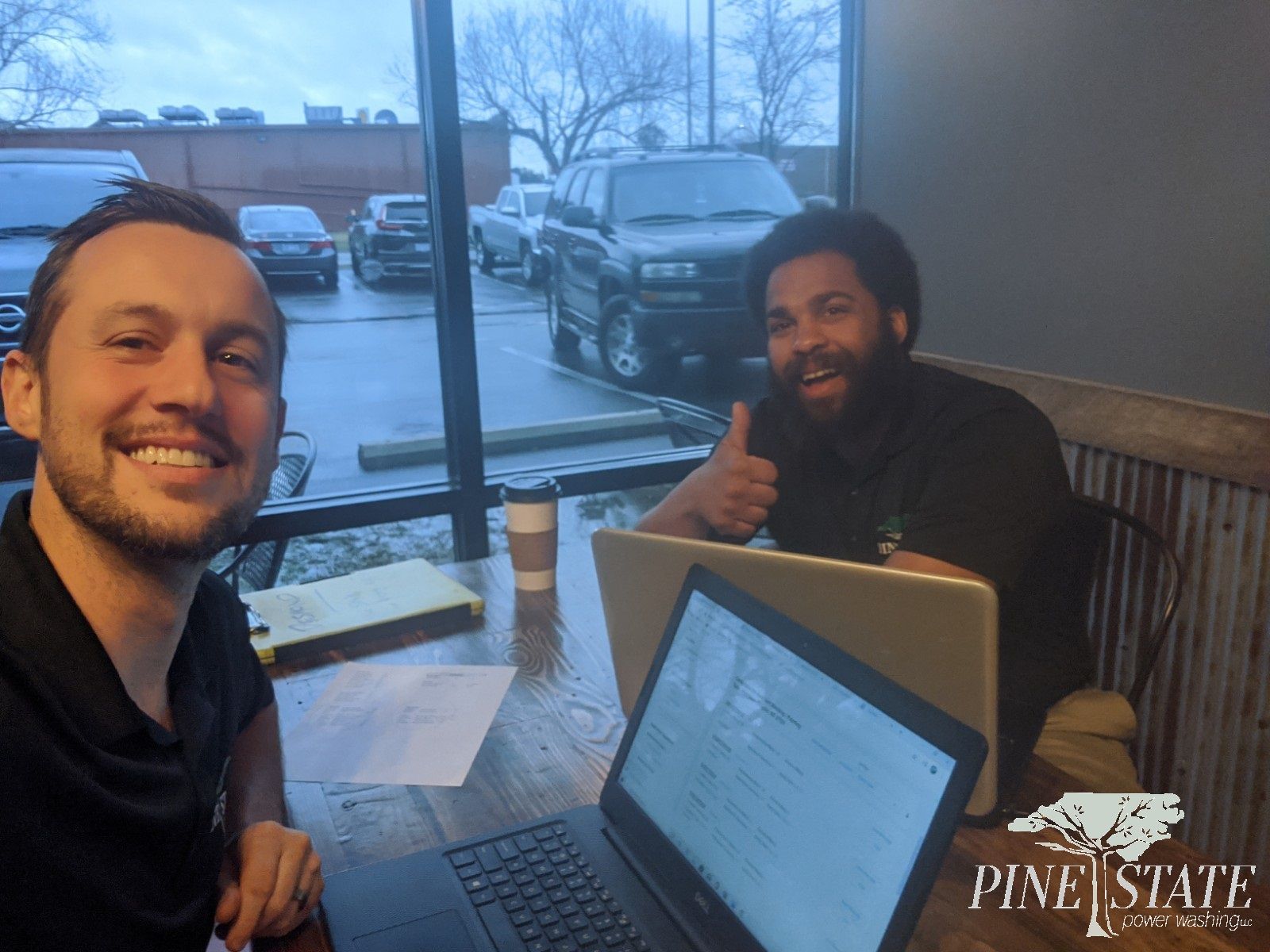 Two men are sitting at a table with laptops and one is giving a thumbs up