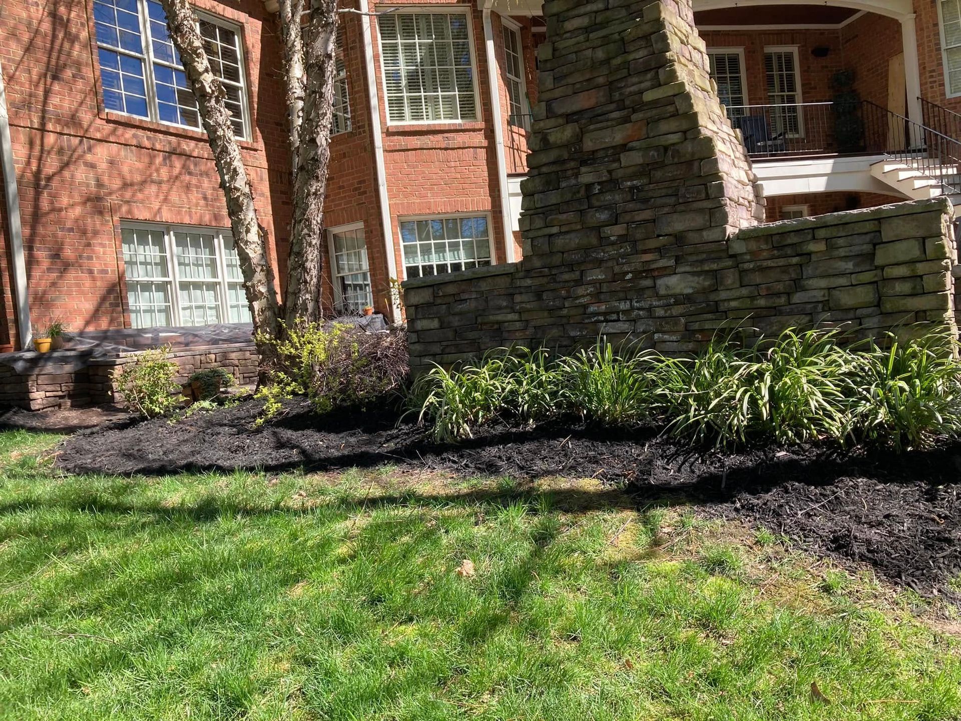 Green lawn with black mulch border in front of brick building and stone structure.
