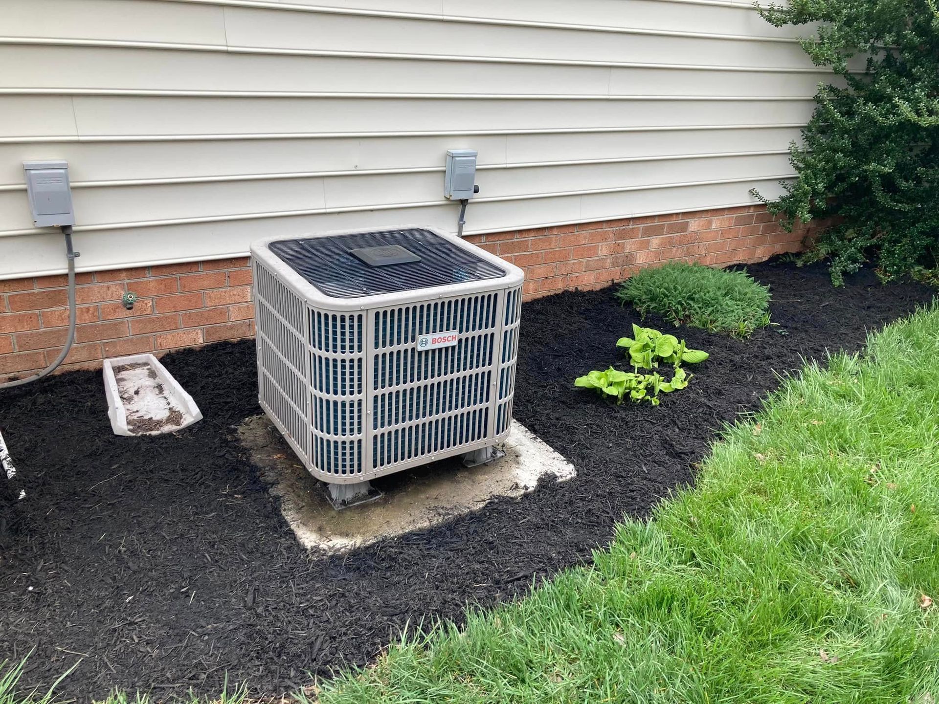 Central air conditioning unit on a concrete pad surrounded by mulch and grass, next to a house.