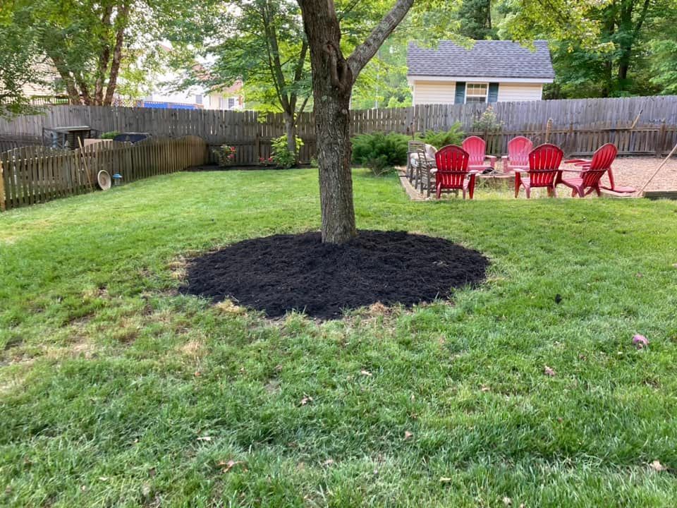 Lawn with tree base mulched in black, red chairs around a fire pit, and a wooden fence.