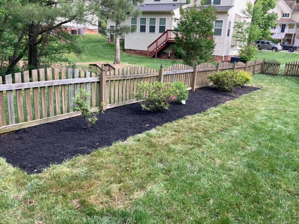 Backyard garden bed with dark mulch and young plants along a wooden fence, green grass.