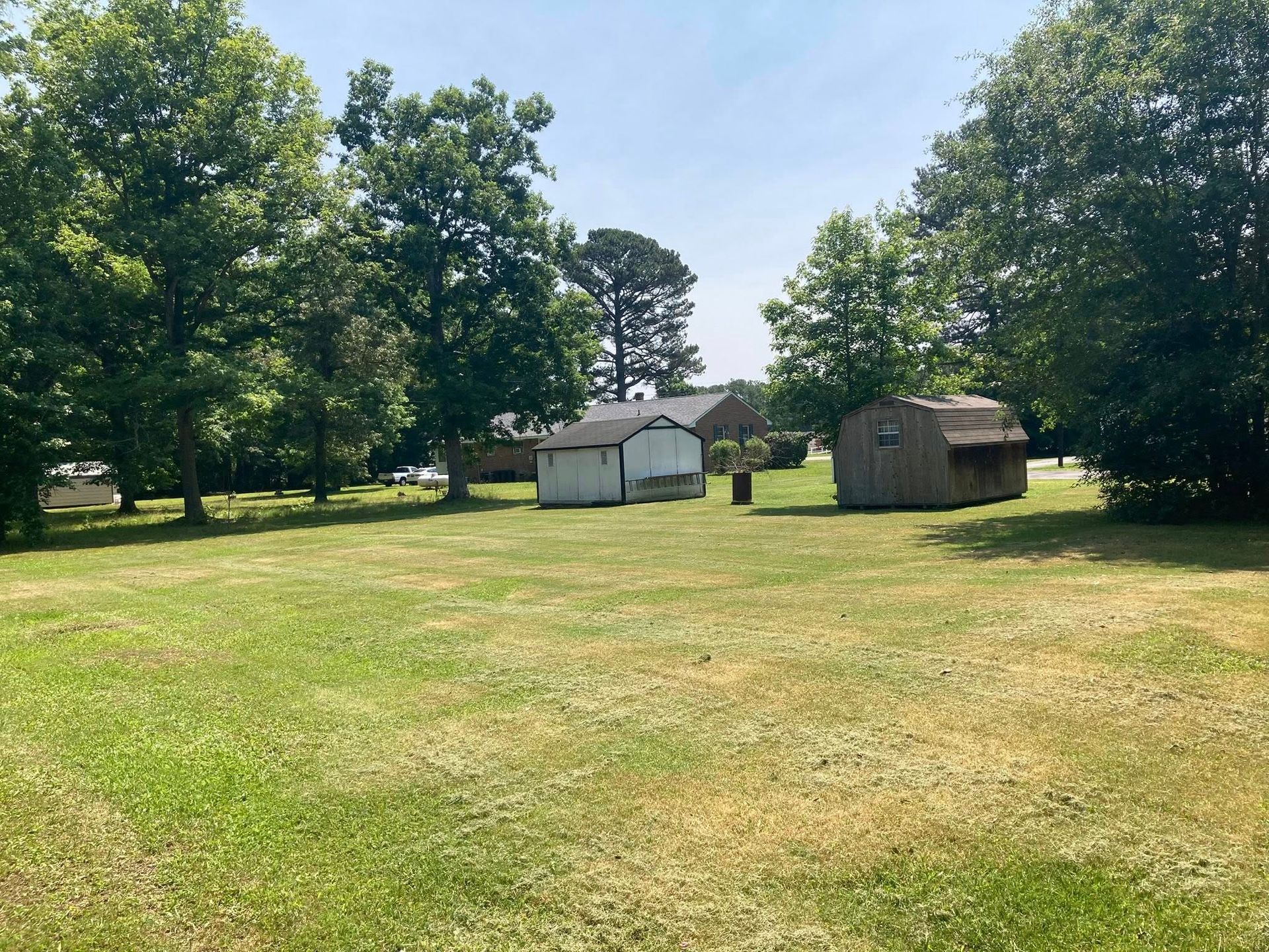 Grassy yard with two sheds, trees, and a house in the background on a sunny day.