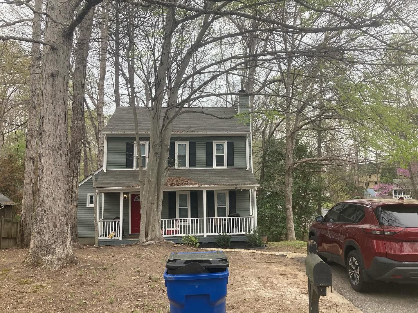 Two-story gray house with white porch and shutters, red door, and trees. Blue trash bin in front.