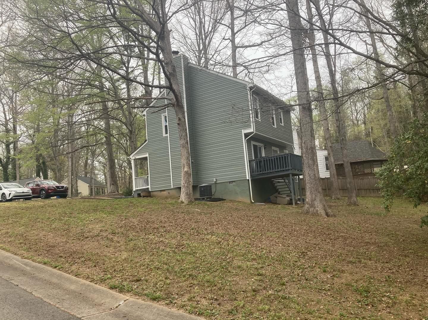 Green house with dark porch on a slight hill, surrounded by trees. Cars parked on the left.