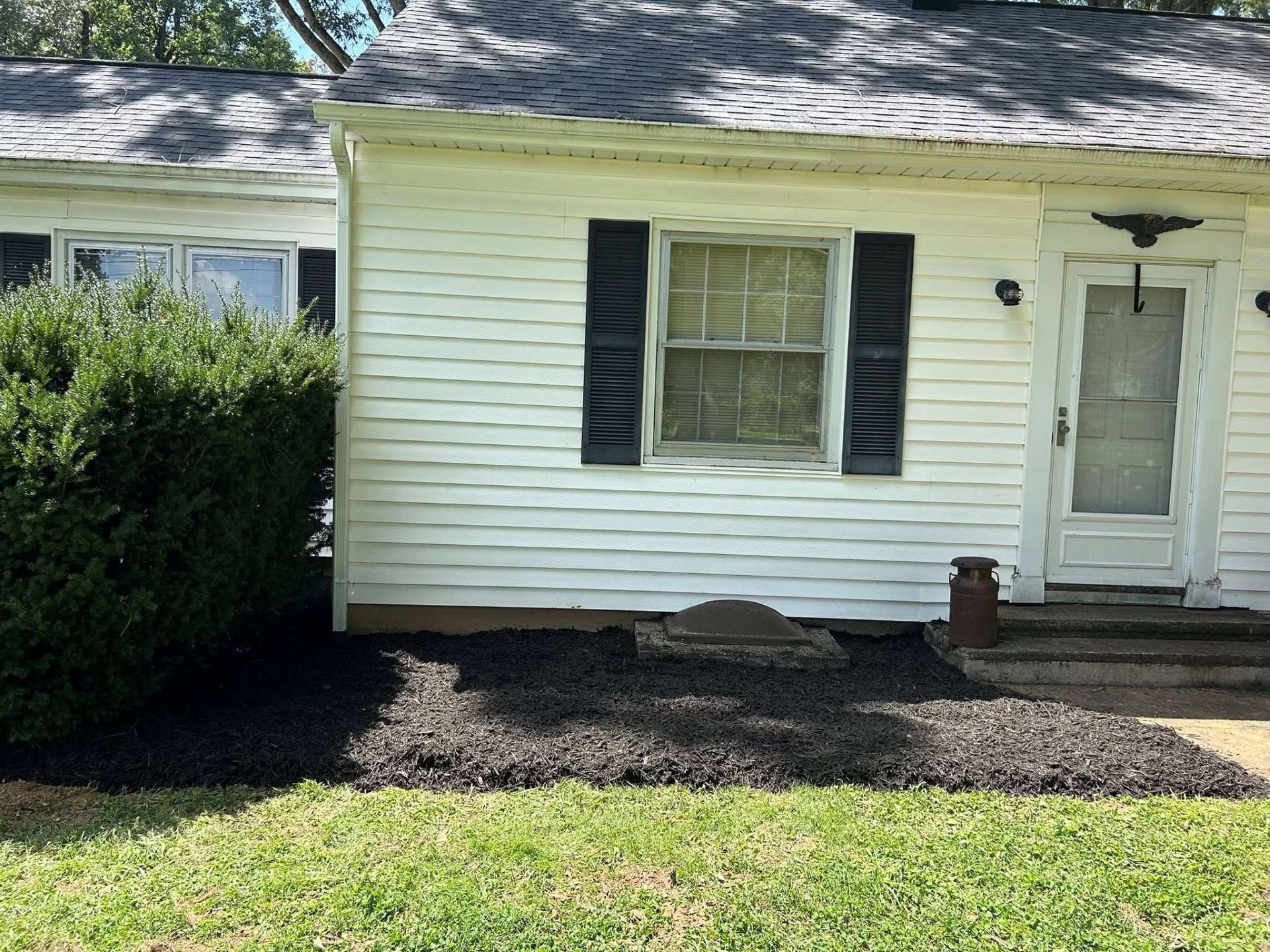 White house exterior with shutters and door, small garden bed with black mulch, green grass.