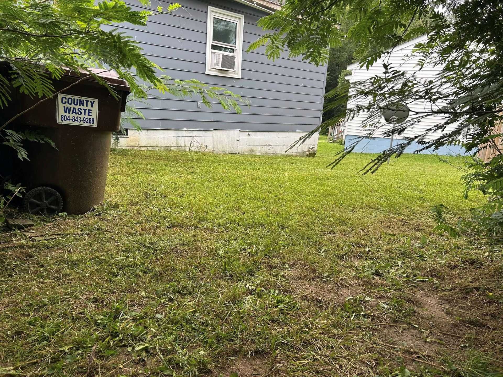 Brown trash can in grassy yard; blue-gray house in background with window AC unit; white building visible.
