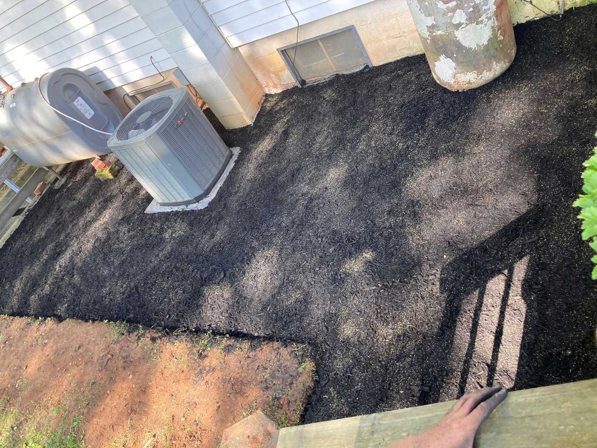 Black mulch covers a backyard area near an air conditioner, building, and concrete objects.
