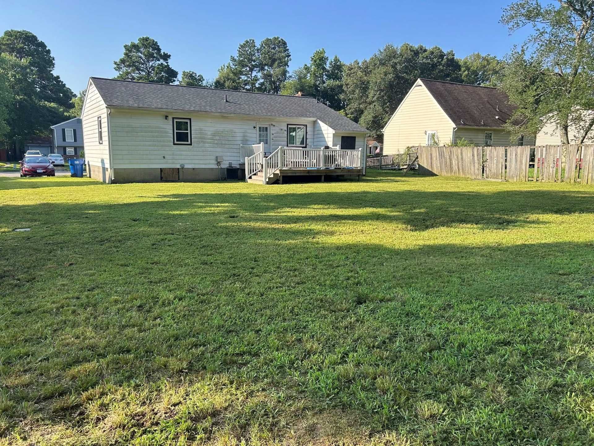 Backyard view of a white house with a deck and a green lawn, surrounded by trees and a wooden fence.