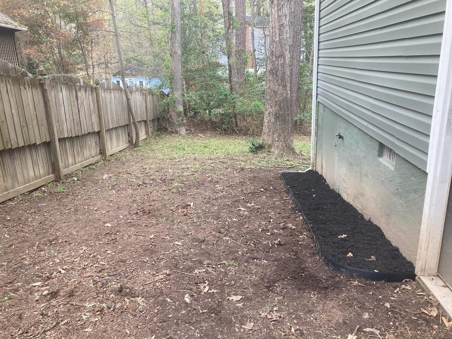 Backyard with a wooden fence, dirt ground, and a house with green siding, mulched area.